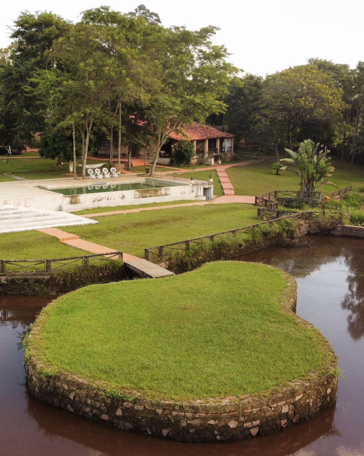 Vista aérea de la laguna con su isla de bordes de piedra, el puente, el jardín y la piscina al fondo