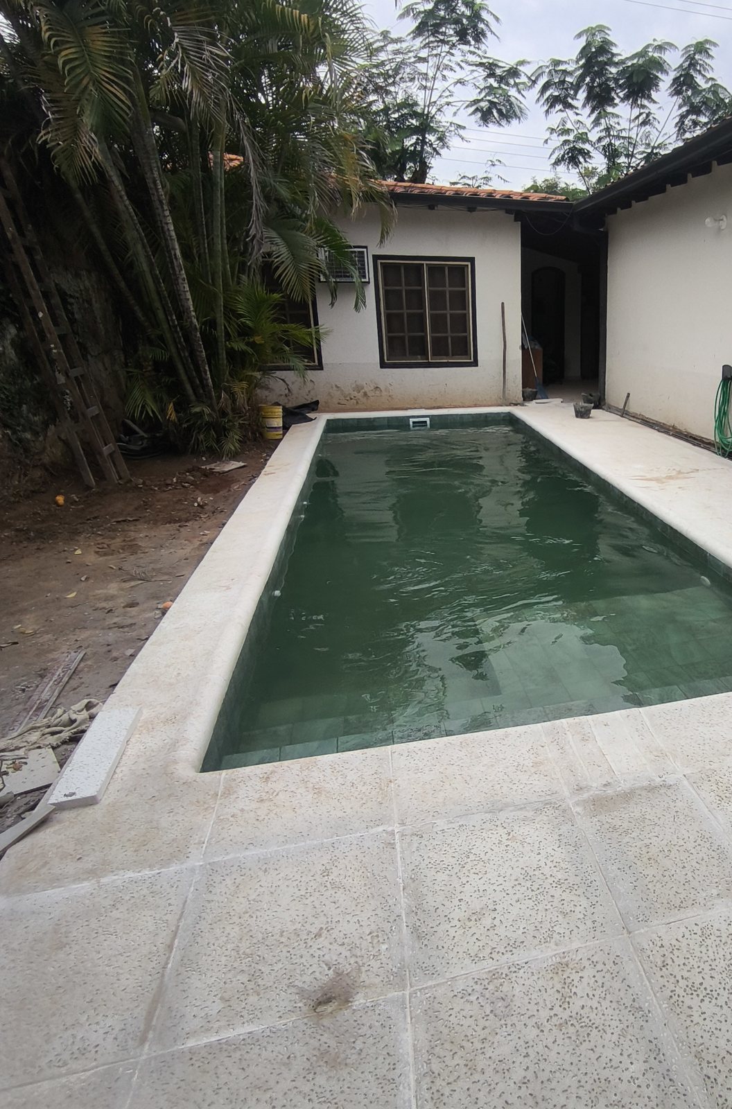 Piscina rectangular con agua, borde y solárium de losetas claras en el patio de una casa